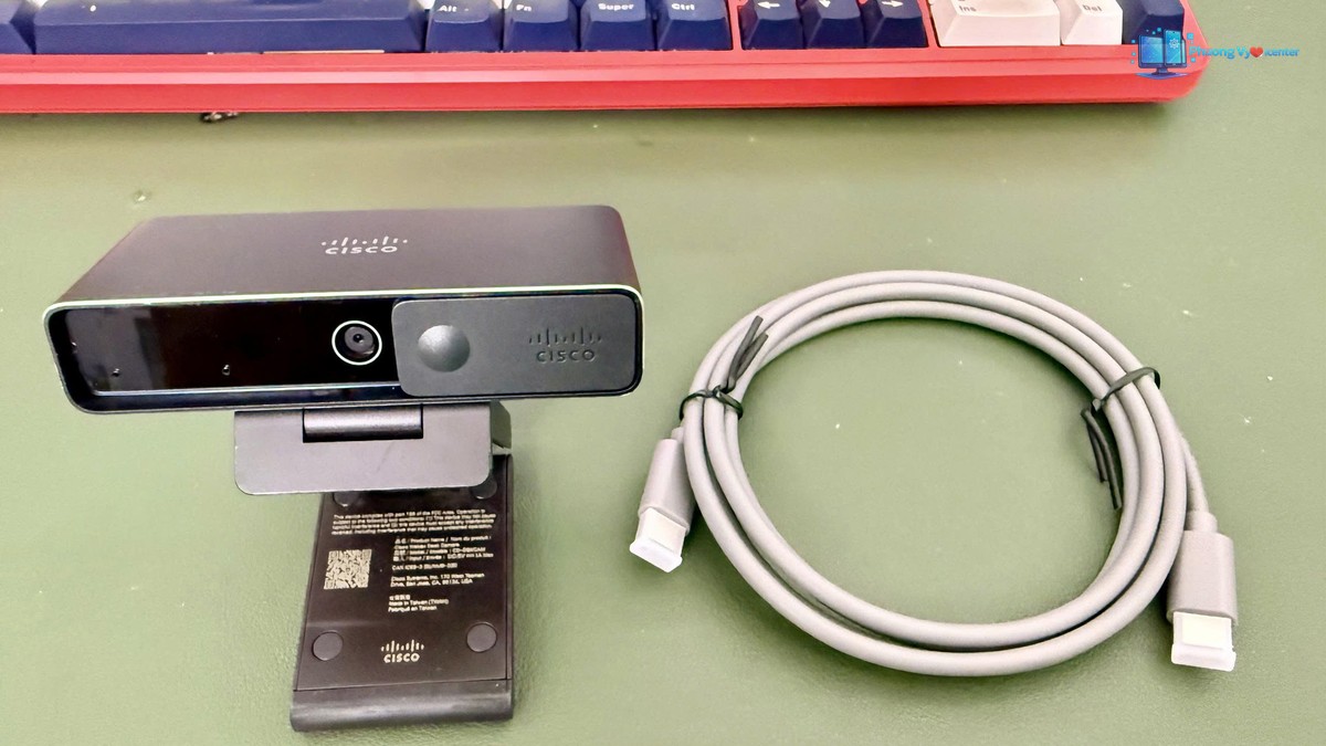 Webcam mở chân kẹp đặt cạnh dây cáp cuộn tròn trên bàn.