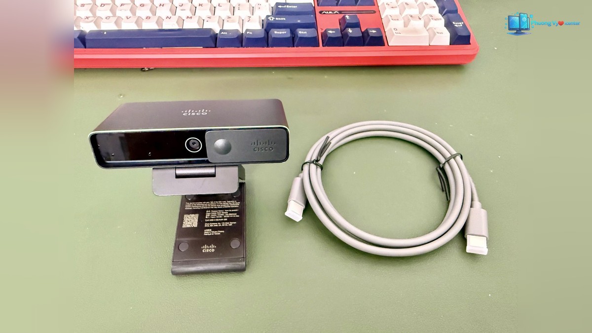 Webcam mở chân kẹp đặt cạnh dây cáp cuộn tròn trên bàn.