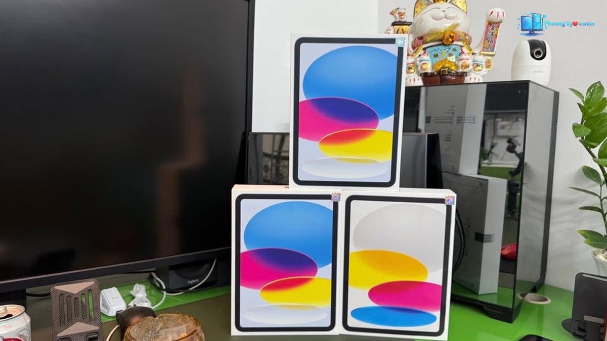 Ba hộp Apple iPad A16 xếp trên bàn trước màn hình và thùng máy tính